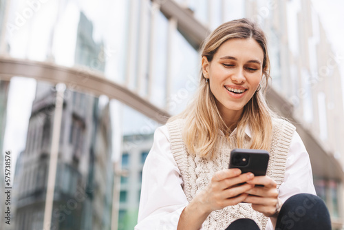 Low angle of glad woman in formal wear sitting in megapolis on street near urban building using smartphone, look happy and smiling. Blonde girl read good news.
