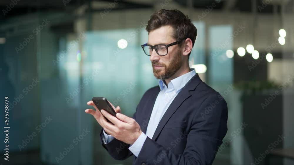 Handsome businessman in formal suit uses mobile phone in modern glass office Bearded male in glasses typing browsing chatting smartphone at workplace Indoor confident entrepreneur financier or broker