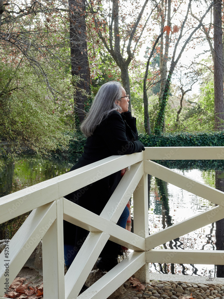 Fototapeta premium Older woman with gray hair and glasses next to a tree feeling it. Woman leaning on the trunk of a tree in a park with green grass. Girl in prairie and green forest pensive and nostalgic