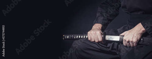 The samurai holding a Japanese katana sword, banner. Photo of a warrior dressed in black clothes in low key with copy space for text