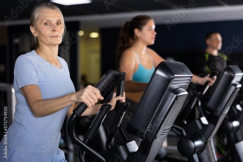 Portrait of motivated sporty senior woman doing cardio training, exercising on elliptical trainer in gym