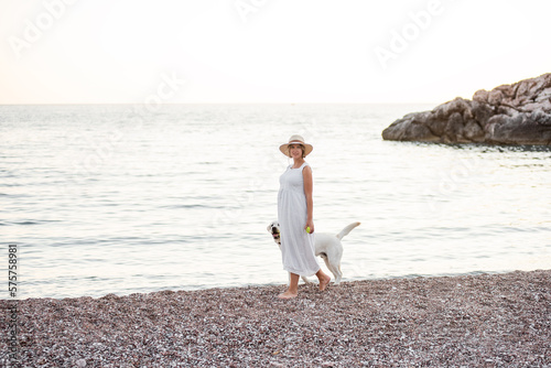 Pregnant future mother walking along the sand of the beach near the sea with golden retriever. Millennial woman in white hat holds dog on leash. Concept of traveling with pets  pet friendly beach