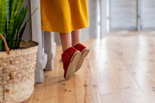 Girl in deep yellow dress is standing on tiptoe by wooden cupboard in the kitchen. Close-up burgundy childs shoes standing on light laminate floor. In handmade rustic planter green flower sansevieria photo