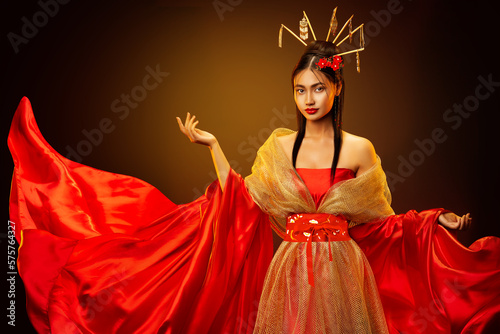 Asian Empress in Red Silk Dress with Golden Jewelry. Beautiful Chinese Girl in Traditional Wedding Costume Clothes. Japanese Model in Fantasy Bride Gown over Dark Studio Background