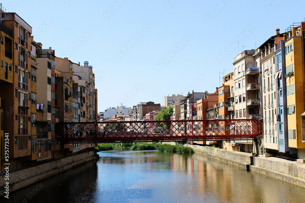 colorful 4- or 5-story buildings border a watercourse, which partially reflects the color of the buildings, with the two banks connected by a red metal bridge