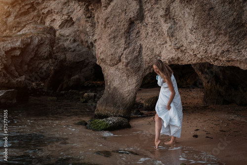 a girl in a white blanket walks along the beach at the sea, near the rocks. Beautiful, slender legs walk along the beach. Cream for varicose veins. High quality photo
