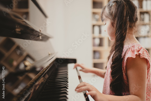 Cute and focused young little Asian girl practicing songs and melody while pressing white and black key and creating new music on piano as hobby and dedication at home