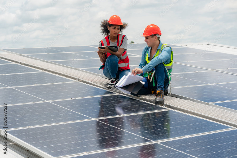 Two young engineer and inspector wearing safety vest and hard helmet inspecting solar panel by kneeling and filling holes and fixing damage using a drill machine while holding a clipboard