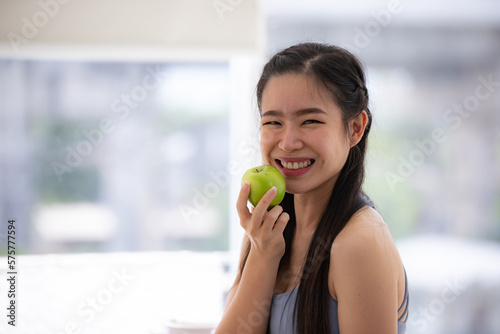 Portrait of healthy and fit young woman sitting on kitchen platform in sportswear holding juicy green apples for weight loss and morning detox drink after workout