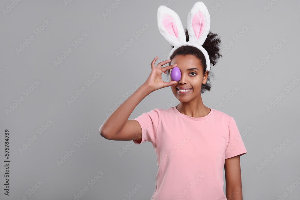 Happy African American woman in bunny ears headband covering eye with Easter egg on gray background. Space for text Happy African American woman in bunny ears headband covering eye with Easter egg on gray background. Space for text