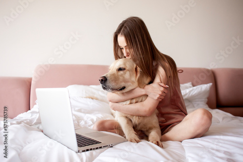 girl lies on bed with golden retriever dog and uses laptop, female freelancer with pet looks at the computer