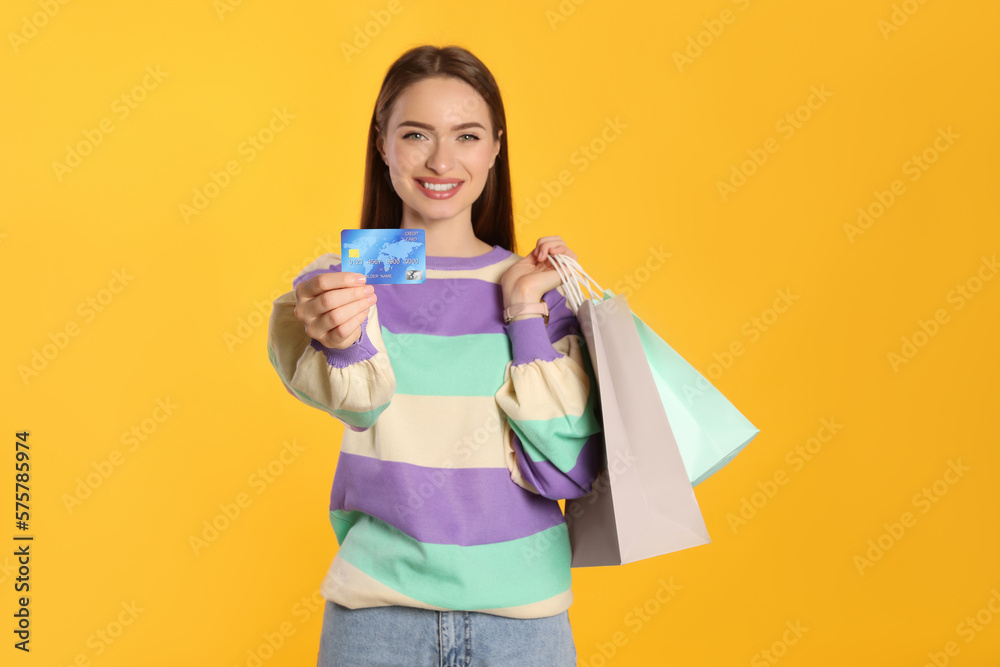 Happy young woman holding shopping bags against yellow background, focus on hand with credit card. Big sale Happy young woman holding shopping bags against yellow background, focus on hand with credit card. Big sale