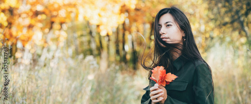 Female beauty portrait surrounded by vivid foliage. Dreamy beautiful girl with long natural black hair on autumn background with colorful leaves in bokeh. Fallen leaves in girl hands in autumn forest.