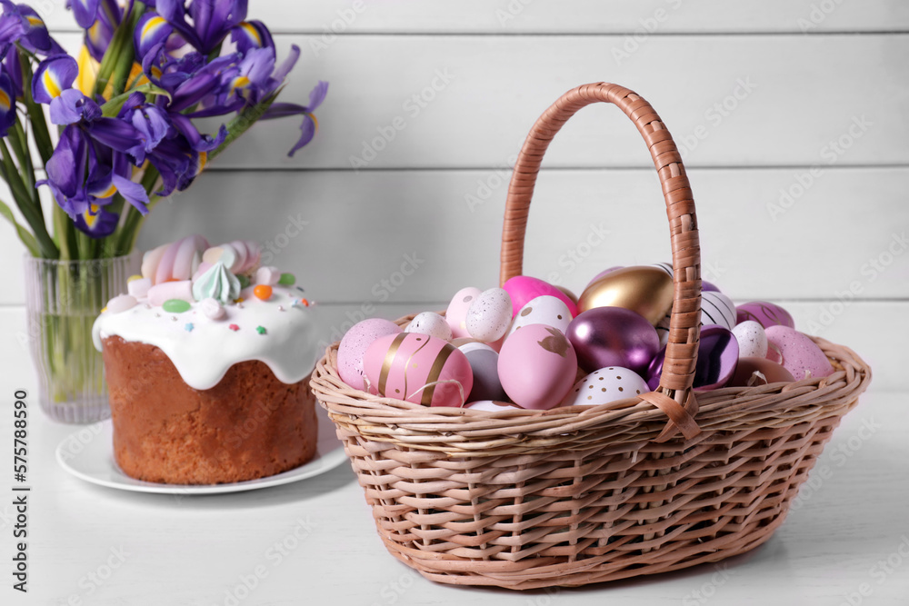Wicker basket with festively decorated eggs, Easter cake and iris flowers on white table