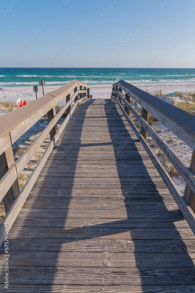 Perspective vertical shot view of wooden walkway with railings at the beach in Destin, Florida. Views of the beach with white sand against blue ocean and sky from the wooden footbridge.