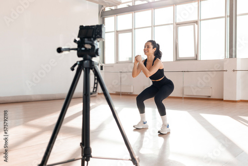 girl in sportswear crouches in the gym and records workout on video camera on tripod, athletic girl sports blogger