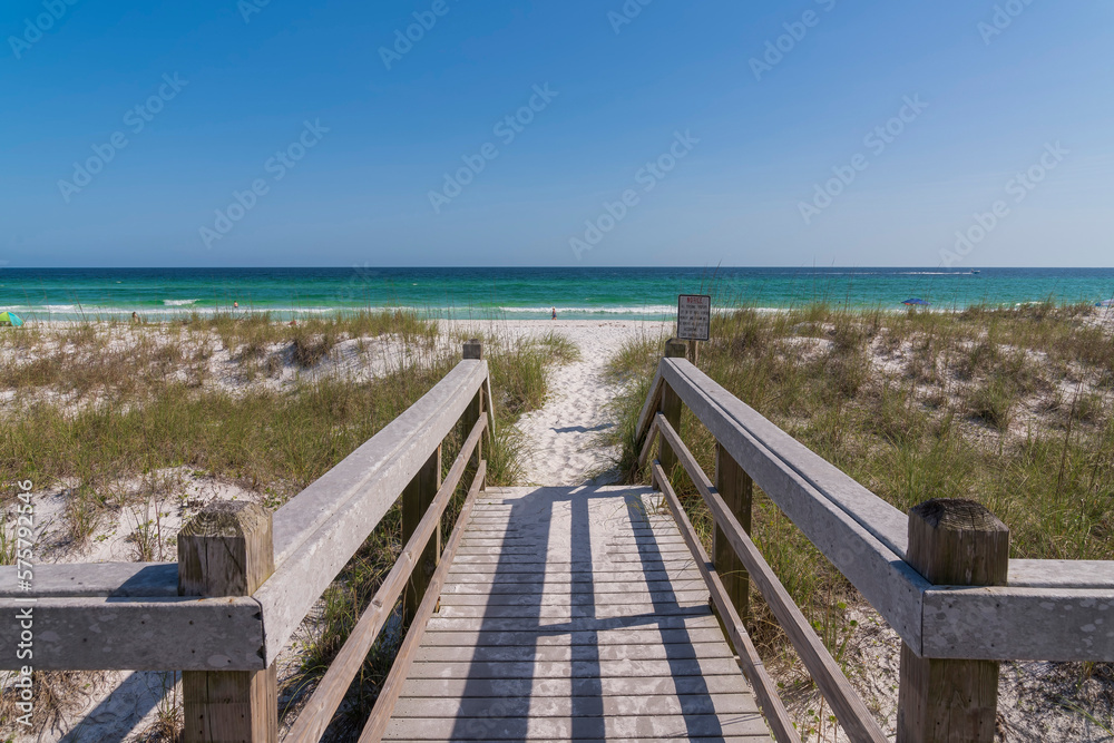 Wooden walkway overlooking ocean and horizon on a sunny day in Destin Florida. The sunlit wooden pathway leads to the sandy shore with view of the clear blue sky.