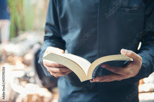 Asian man holding book reading magazine at green park in natural garden. Young man relaxation read open book self study. University men happy learning. Closeup Men hands open textbook in green park © aFotostock