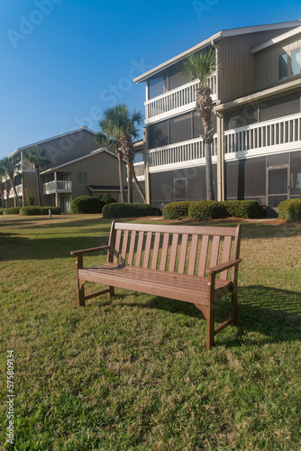 Wooden bench facing front near the townhomes buildings behind in Destin, Florida. Bench on a lawn at the front of townhouses with screen walls and palm trees outdoors.