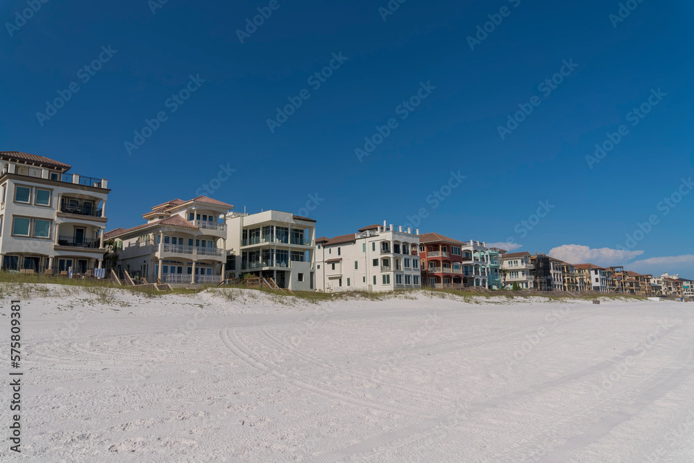 White sand at the front of beach houses with footbridges over sand dunes in Destin, Florida. Row of houses with balconies and roof decks against the blue sky background.