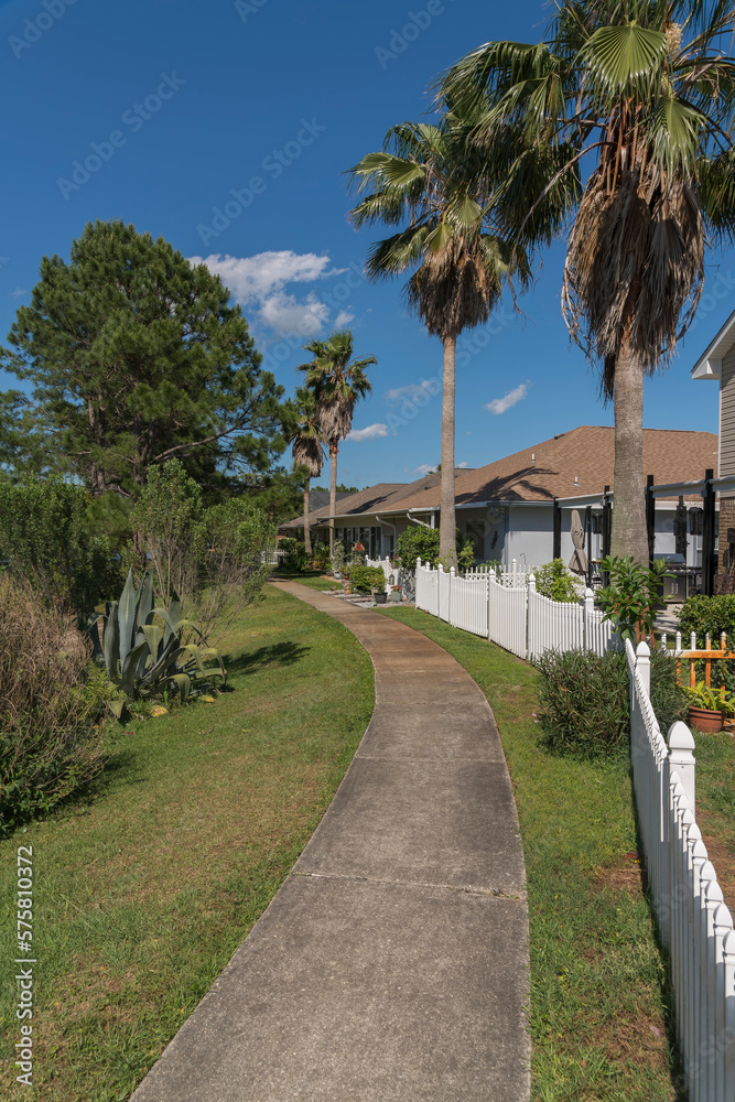 Curved concrete walkway with bushes on the side outside the fenced residences in Navarre, Florida. Houses with palm trees inside their yard behind the picket fence near the curved path in the middle.