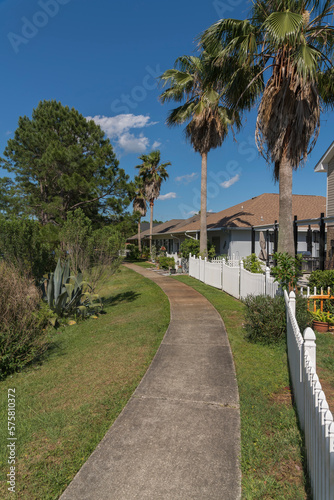 Curved concrete walkway with bushes on the side outside the fenced residences in Navarre, Florida. Houses with palm trees inside their yard behind the picket fence near the curved path in the middle. © Jason