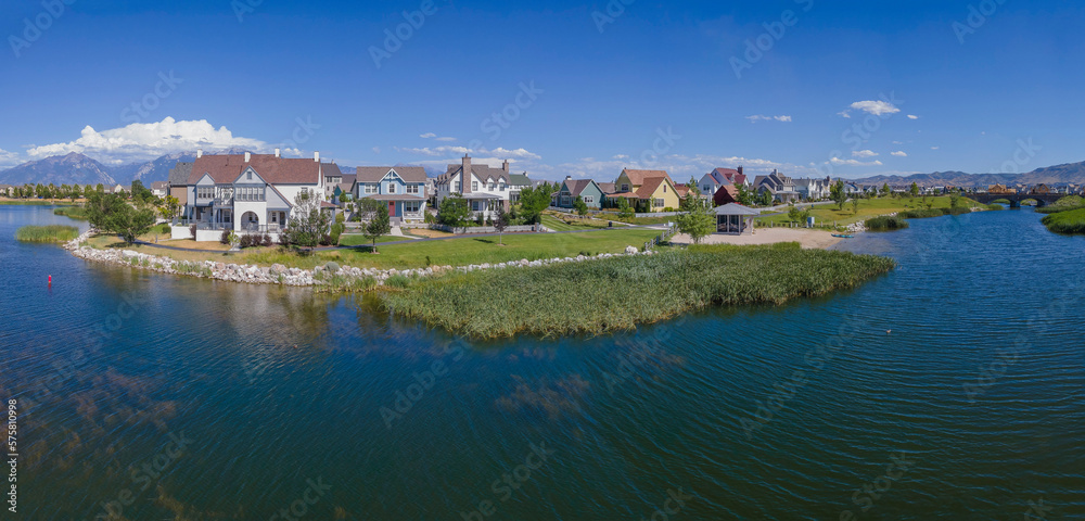 Aerial view of lakefront single-family homes against the blue sky at Daybreak, Utah panorama. Lake with grasses near the shore and view of mountains at the background.