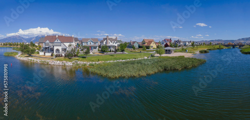 Aerial view of lakefront single-family homes against the blue sky at Daybreak, Utah panorama. Lake with grasses near the shore and view of mountains at the background.