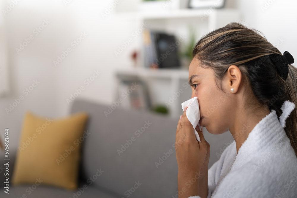 Young lady, sitting on the couch in the living room, while blowing her nose Young lady, sitting on the couch in the living room, while blowing her nose