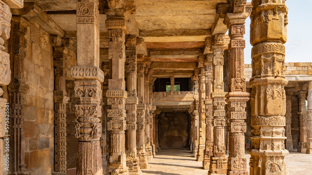 Quwwat-ul-Islam mosque in the Qutub Minar temple complex. The gallery with elegant carved sandstone columns goes forward. Green vegetation is visible through the window. Blue sky. India. Delhi