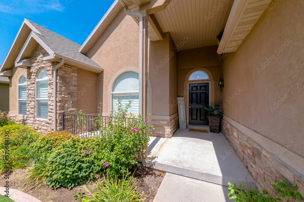 Obraz premium Entrance of a house with painted light brown stucco and stone veneer wall. House with plants near the arched windows on the left and black front door with transom and panel and plants on the right.