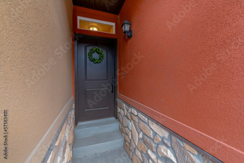 Black front door with wreath, transom window, and concrete steps at the entrance. House entrance in between the light brown stucco wall on the left and orange stucco wall on the right.