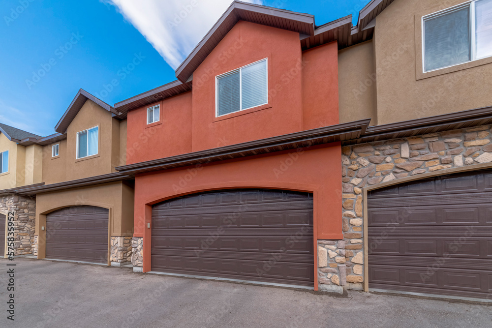 Townhouses with attached garages and painted orange and brown stucco and stone veneer walls. There are brown garage doors with arched entrances of two-storey townhomes under the clear sky.