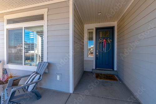 Dark blue front door with sidelight, wreath , and lockbox. House with gray wood lap siding and lounge chairs near the window with reflective glass panes at the entrance.