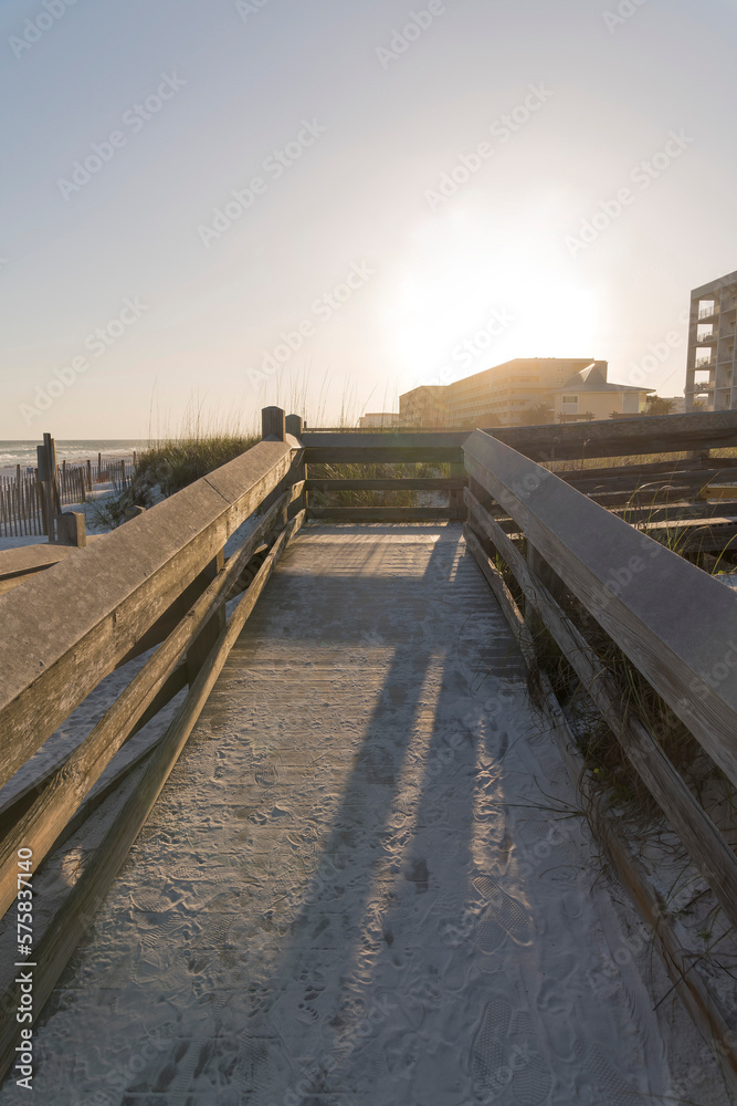 Sand covered wooden pathway with sunset views behind the buildings in Destin, Florida. Sloped pathway with railings near the sand dunes at the beach.
