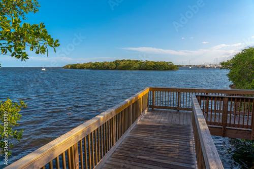 Boardwalk corner with railings and views of an island near the Dinner Key Marina at Miami, Florida. Corner of a wooden path near the trees with views of the water and island against the blue sky. © Jason