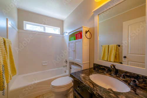 Small bathroom interior with marble sink counter  toilet shelves  and acrylic tub shower. There is a sink on the left beside the toilet with wooden cabinet above across the hanging two yellow towels.