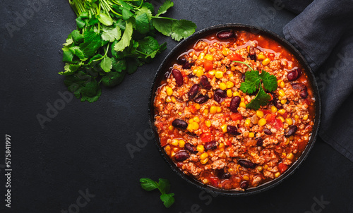 Chili con carne with beef, red beans, paprika, corn and hot peppers in tomato sauce, spicy tex-mex dish in a cooking pot, black table background, top view