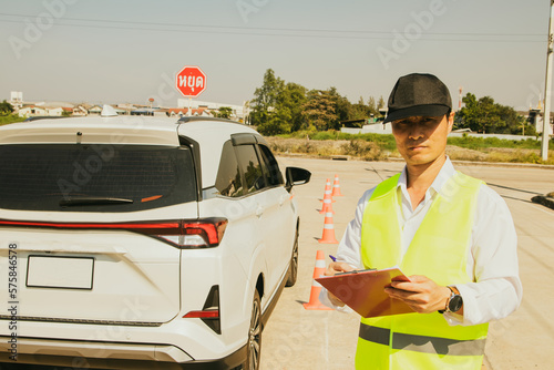 Driving test concept : Asian male officer in charge of legal driving license exam holding clipboard and test papers prepared and earnestly stands dignified on the road of the field test field. photo