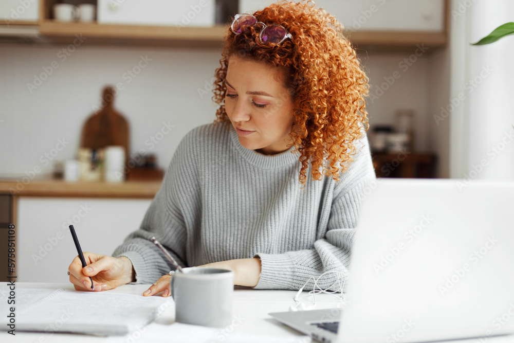Redhead female interpreter freelancer with curly hair working from home, writing translation in her copybook sitting at kitchen table in front of laptop, translating article for web site - obrazy, fototapety, plakaty 