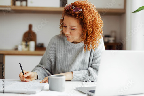 Redhead female interpreter freelancer with curly hair working from home, writing translation in her copybook sitting at kitchen table in front of laptop, translating article for web site