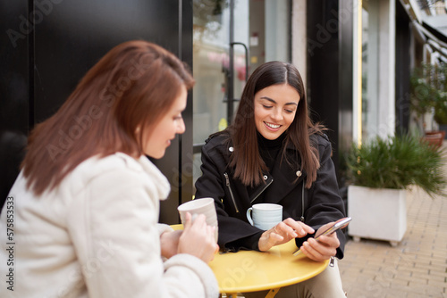 Female friends smiling while using a mobile phone sitting at coffee shop outdoors. Technology and friendship concept.
