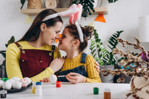 Caring young mother hugging happy pretty little daughter child, funny family painting eggs sitting at table, wears bunny ears. Easter celebration and traditions. Concept creativity and spring holiday