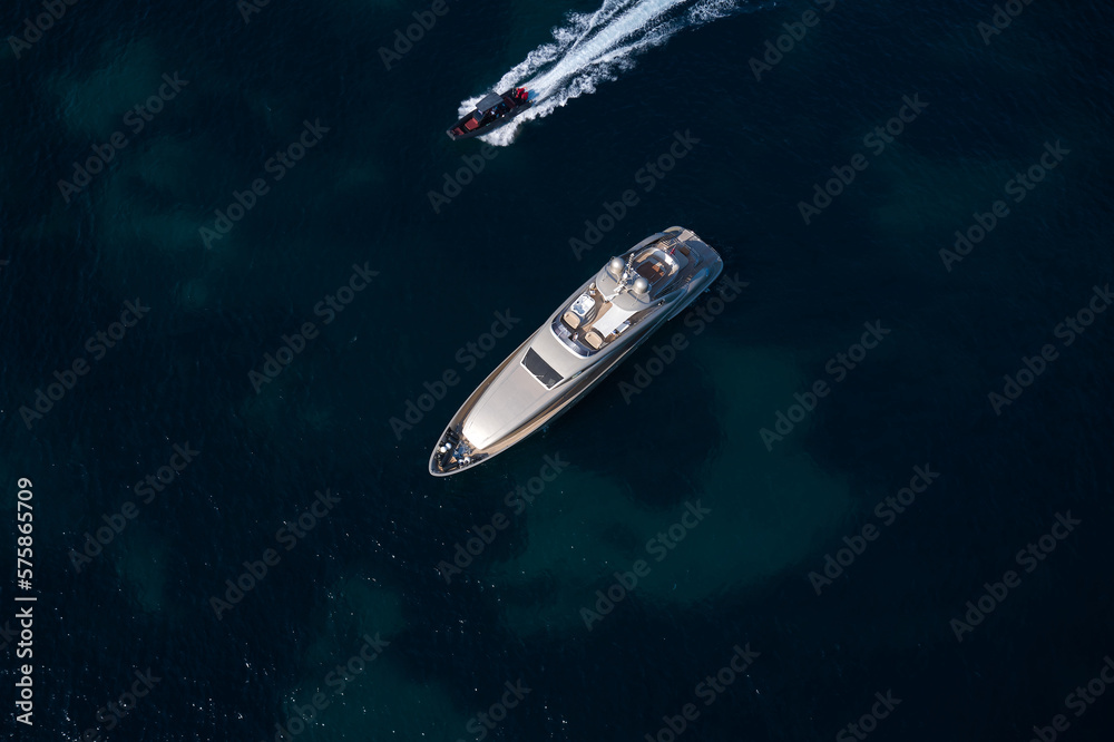Large open yacht anchored in the blue sea top view. Silver modern boat on blue transparent water aerial view. Boat fast moving next to yacht top view. Large open yacht anchored in the blue sea top view. Silver modern boat on blue transparent water aerial view. Boat fast moving next to yacht top view.