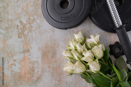 Gym dumbbells barbell weight plates and white roses flowers. Gift for Women's Day, Mother's Day, birthday, wedding anniversary or marriage proposal engagement. Fitness workout flat lay with copy space