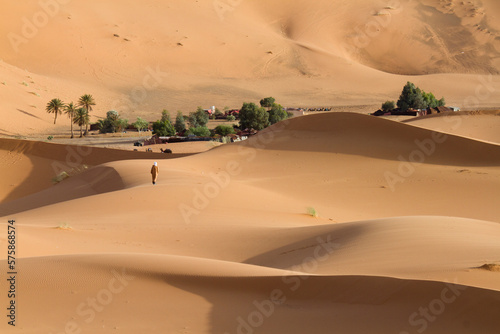 Moroccan man wearing the traditional Djellaba walks in Red sand dunes in Erg Chebbi towards a camping site, Sahara desert. Merzouga, Morocco