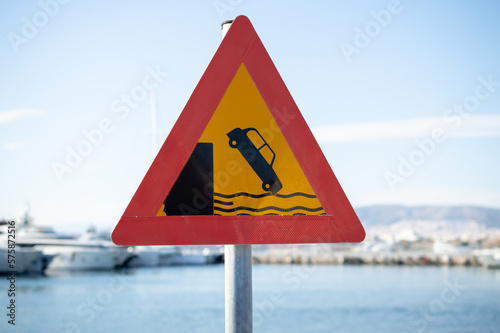 Triangular warning road sign, yellow with red outline depicting a car falling off a harbor dock. Drive carefully, pier embankment