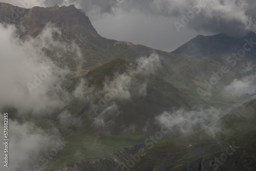 Stormy dramatic weather in mountain with fluffy grey clouds above high rocky ridges with green slopes and deep gorge in summer. Mountain landscape in bad weather.