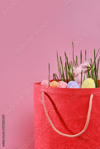 Red shopping bag, green grass and Easter eggs on pink background with copy space. Online shopping for the holidays. Symbols, traditions concept.
