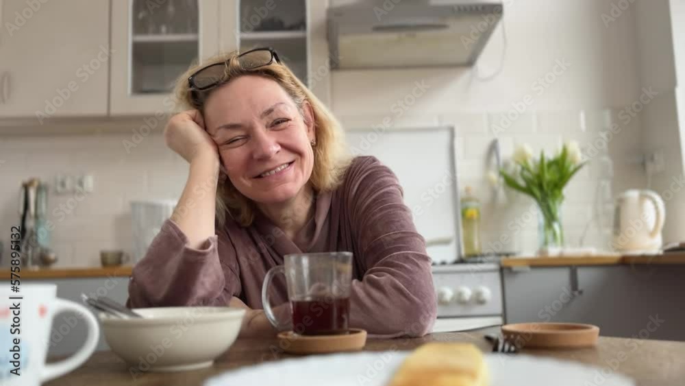A middle-aged woman in glasses drinks coffee for breakfast sitting at the table in the kitchen sincerely laughs. Emotions A middle-aged woman in glasses drinks coffee for breakfast sitting at the table in the kitchen sincerely laughs. Emotions